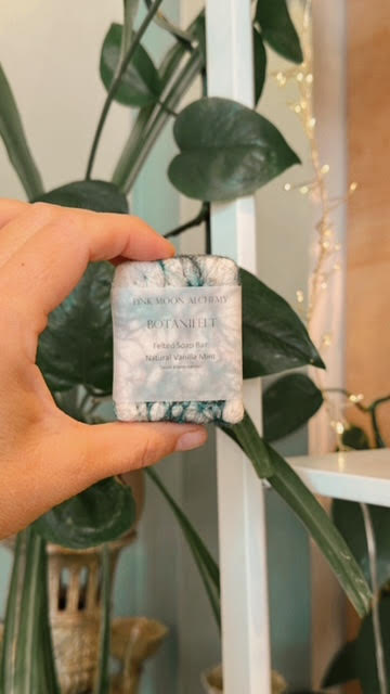 Hand holding a natural felted soap with plants in the background