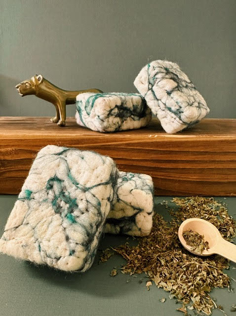 Three white and green felted soap bars on a wooden block with dried herbs and a wooden spoon on a green surface. Felted Natural Soap
