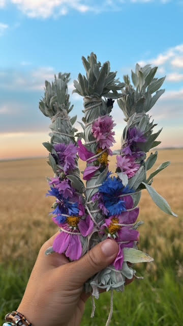 Hand holding a bouquet of purple and blue flowers against a natural background. A Sage Wand